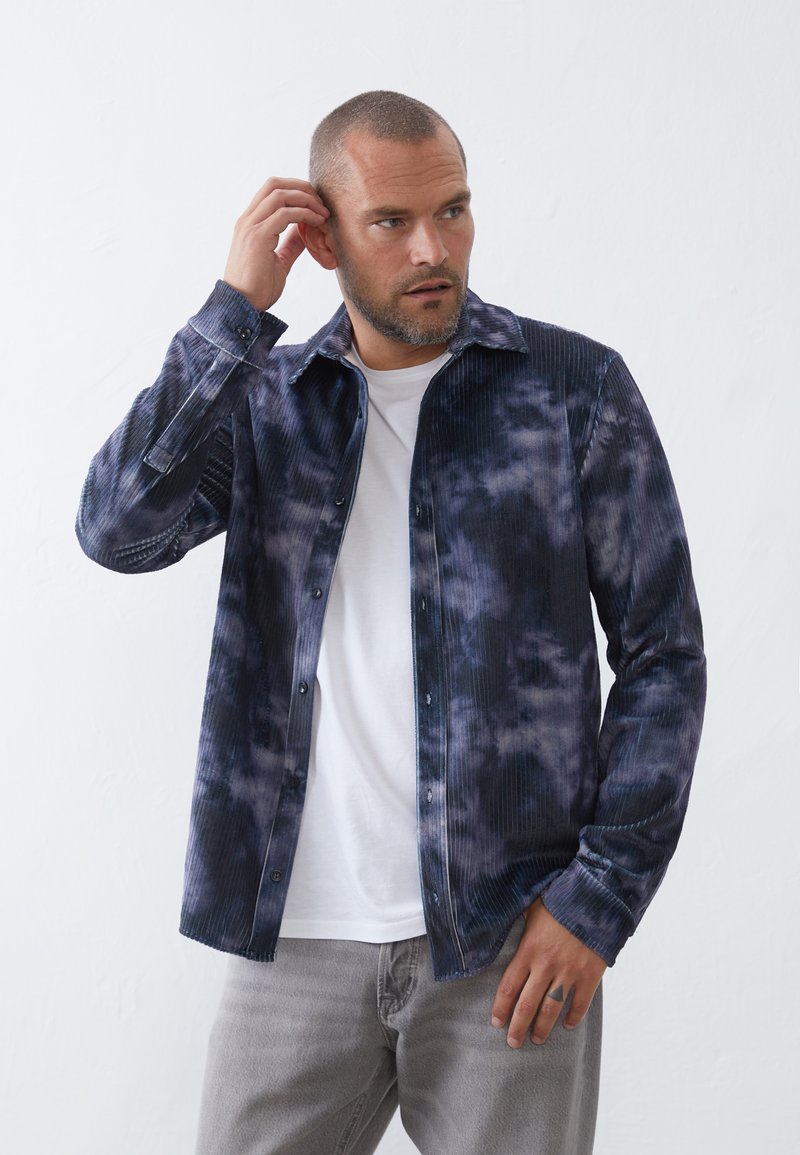 Camisa de corduroy en color azul marino con efecto tie-dye, con costillas verticales, cierre de botones en la parte delantera y cuello puntiagudo, superpuesta sobre una camiseta blanca.