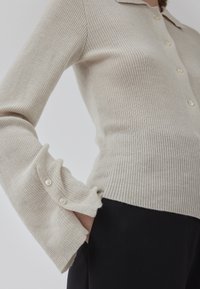 Cardigan beige en maille côtelée avec un col, doté de poignets boutonnés et d'une coupe ajustée. Fabriqué en matériau textile doux.