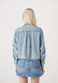 Jaqueta de ganga em tecido azul claro com mangas longas, punhos com botão e um design cropped. Apresenta uma costura em yoke e um ajuste solto.