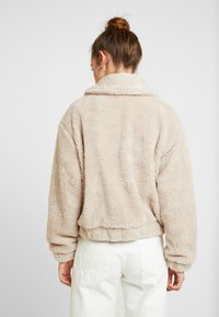 Beige, fluffig, kort jakke med krage och långa ärmar. Texturen liknar mjuk fleece med en avslappnad passform. Bärs över vita byxor.