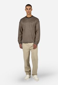 Barna, texturált, hosszú ujjú pulóver, kerek nyakkivágással, bézs nadrággal és fehér sportcipővel párosítva, egy egyszínű háttér előtt állva.