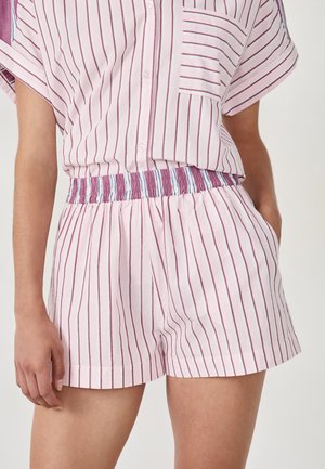 Femme portant une chemise à boutons rayée rose et blanche ainsi que des shorts élastiques assortis, avec une main dans la poche.