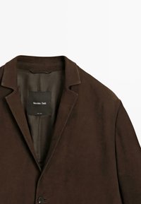 Bruine blazer met een gladde textuur, enkele knoopsluiting en notch-lapels. Interieur heeft een contrasterende voering met een label.