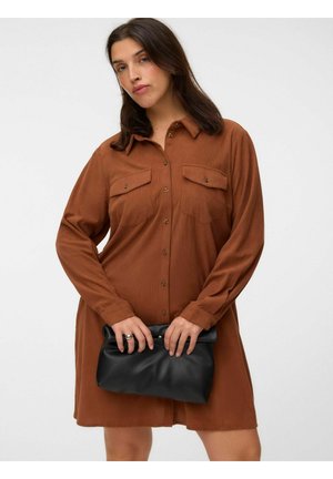 Kvinde iført en brun kjole med knapper, der holder en sort læderclutch med begge hænder foran en ensartet lys baggrund.