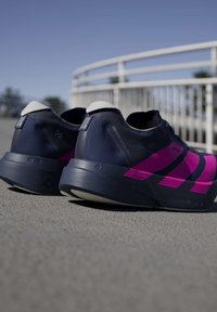 Paire de chaussures de course bleu marine et rose placées sur un trottoir près d'une rambarde métallique blanche sous un ciel dégagé.