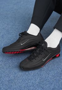Črne športne čevlje z teksturirano zgornjico, ki vsebujejo izrazit swoosh in rdeče podplate. Ustrezno kombinirani z belimi nogavicami in črnimi hlačami.