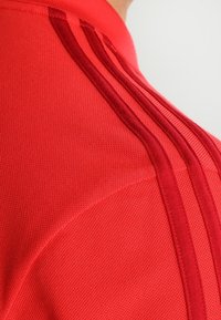 Polo de sport rouge avec trois rayures verticales de la même teinte. La texture est lisse avec un léger côtelé au niveau du col.