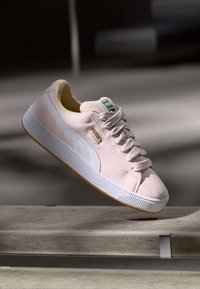 Baskets Puma en suède rose clair avec bande blanche, semelle blanche et logo Puma doré, flotter au-dessus d'une surface en béton.