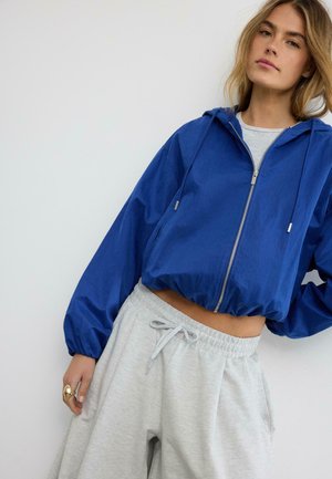 Femme portant un sweat à capuche court bleu avec fermeture éclair et un pantalon de survêtement gris clair à cordon, posant devant un fond blanc.