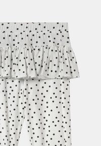 Biała tkanina w czarne kropki z detalem w postaci marszczonej falbany w górnej części, widoczna elastyczna talia.