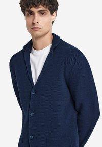 Mörkblå stickad cardigan med ett strukturerat mönster, sjalkrage, fem knappar och två framfickor. Bärs över en vit skjorta.