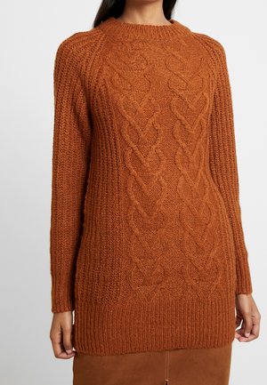 Rustfarvet kabelstrikket trøje med lange ærmer og rund hals, båret af en person med mørkt hår.