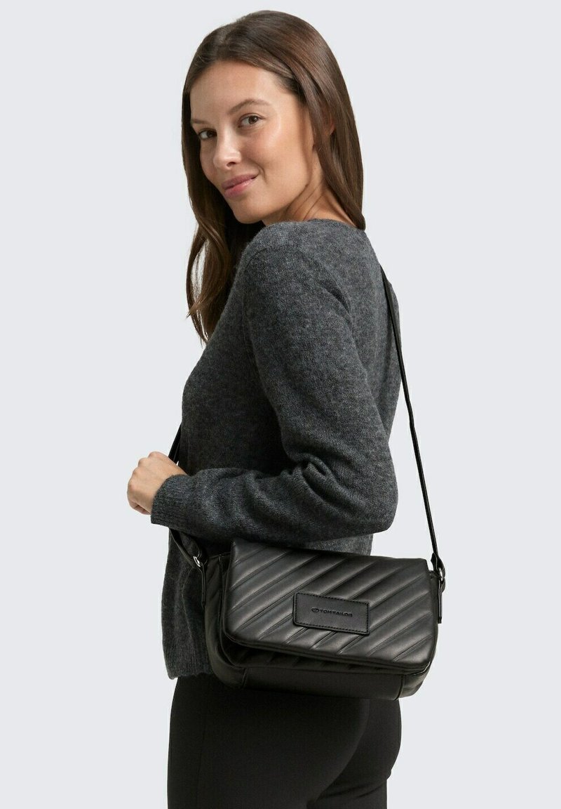 Bolso cruzado acolchado negro fabricado con material sintético, con cierre de solapa y correa ajustable, que cuenta con un parche de logo en la parte frontal.