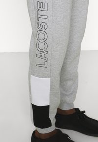Šedé tepláky s černým logem "LACOSTE", bílo-černé barevné bloky na spodní části nohavice, vyrobené z měkké tkaniny. Boty jsou černé.