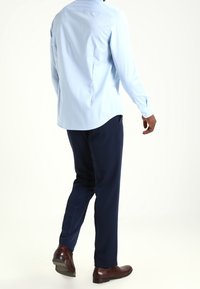 Ljusblå, långärmad skjorta med knäppmanchetter, ihop med marinblå byxor och bruna läderskor, sedd bakifrån.