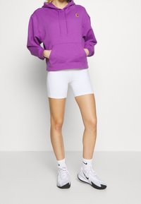 Felpa in cotone viola con tasca frontale e cappuccio con cordoncino, abbinata a pantaloni bike bianchi e scarpe da ginnastica bianche con dettagli rosa.