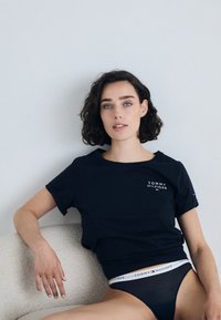 Juodas trumpomis rankovėmis medvilninis marškinėliai su apvalia iškirpte ir balta "TOMMY HILFIGER" logotipu virš juosmens, saugiai derinami su mėlynomis apatinėmis kelnaitėmis su logotipų juostele.