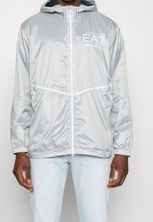 Mann trägt eine silbergraue Kapuzenjacke mit Reißverschluss und dem Logo „EA7 Emporio Armani“ sowie hellblaue Jeans, steht vor einem einfarbigen Hintergrund.