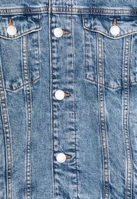 Veste en denim, couleur bleu clair, avec des boutons-pression argentés, deux poches poitrine, des coutures verticales et un léger effet de décoloration pour la texture.