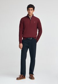 Sudadera de punto texturizado en color marrón, pantalones azul marino, zapatos marrones de cordones con suelas de goma, de pie contra un fondo neutro.