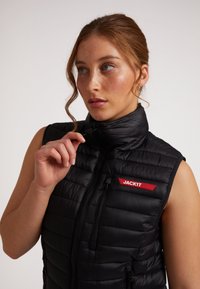 Gilet imbottito nero con colletto alto, chiusura a zip e tasche laterali. Presenta il marchio rosso "JACKIT". Materiale leggero e con texture.