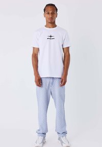 Camiseta blanca de cuello redondo con logo negro, combinada con pantalones jogger azul claro con estampado. El modelo lleva zapatillas blancas y grises.