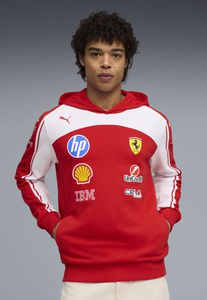 Jeune homme portant un sweat à capuche Ferrari rouge et blanc avec des logos de sponsors tels que HP, Shell, IBM, UniCredit et CEVA Logistics, les mains dans les poches.
