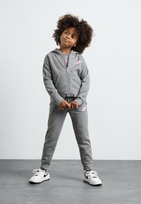 Sudadera con cremallera gris y pantalones de chándal a juego con el logo de Nike en rosa, fabricados en un tejido suave. Zapatillas blancas con toques negros completan el conjunto.