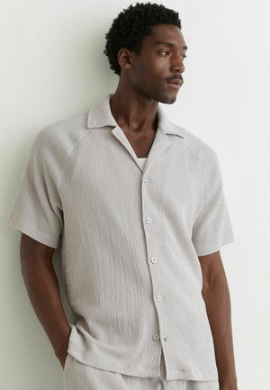 Muž s krátkymi kučeravými vlasmi v svetlo béžovej textúrovanej krátkorezej košeli na gombíky, stojaci oproti bielej stene a pozerajúci do strany.