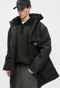 Must musta jope, millel on karvane kapuuts, kantud tekstuuriga halli kudumi sviitri peal, koos hallide triibuliste pükstega ja tume müts.