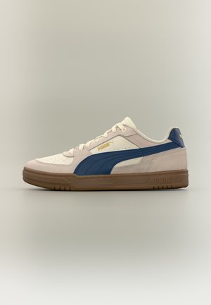 Niskie sneakersy Puma z beżową i kremową cholewką, ciemnoniebieskim paskiem po bokach, białymi sznurowadłami i brązową gumową podeszwą na neutralnym tle.