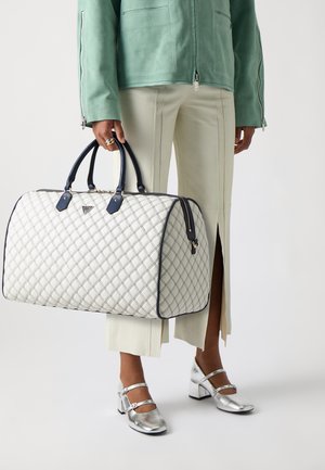 Borsa bianca trapuntata con accenti blu navy, forma arrotondata e manici doppi. Presenta una chiusura con zip e una finitura lucida, posizionata su pantaloni color crema.