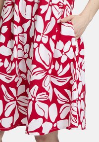 Rotes Kleid mit einem auffälligen weißen Blumenmuster, aus leichtem Stoff gefertigt. Verfügt über seitliche Taschen und ein gerafftes Taillendesign.
