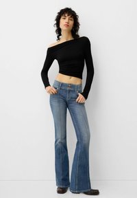Schwarzes Langarm-Oberteil mit schulterfreiem Schnitt, kombiniert mit hellblauen Schlagjeans, die einen Zwei-Knopf-Bund und Vordernähte aufweisen. Glattes, dehnbares Material.