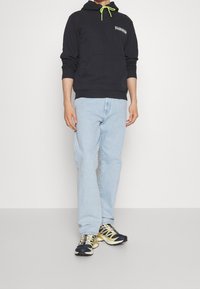 Sudadera con capucha azul marino con cordón de neón verde y bolsillo frontal. Jeans azul claro de pierna recta y zapatillas deportivas negras con amarillo.