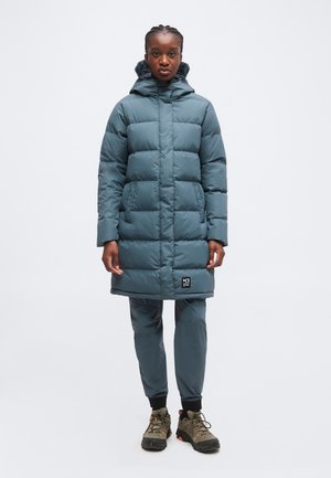 Femme debout portant un manteau d'hiver matelassé à capuche turquoise, un pantalon assorti et des chaussures de randonnée foncées sur un fond clair uni.