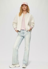 Lichtbeige bomberjack, roze pet met rood logo, roze top, vervaagde blauwe jeans en witte sneakers. Model staat met handen in de zakken.