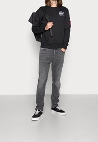 Sudadera negra con logo de la NASA, jeans grises y zapatillas negras con detalles blancos. El modelo lleva un bolso negro con una correa al hombro.