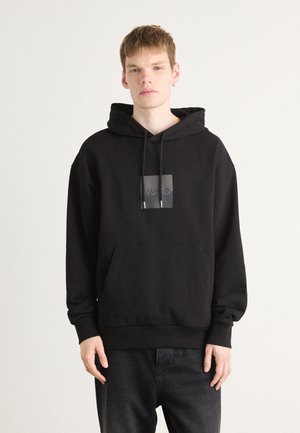 Svart hettegenser med frontlomme, justerbar hette med snor og solid svart firkantet logo på brystet. Brukt med mørke jeans.