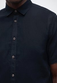 Κοντινό πλάνο ενός ατόμου που φοράει κοντομάνικο πουκάμισο σκούρου ναυτικού χρώματος με γιακά, πάνω σε μονόχρωμο φόντο.