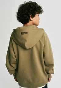Oliwkowa bluza z kapturem o luźnym kroju, z teksturowanej tkaniny i czarnym logo na plecach. Rękawy i dół wykończone ściągaczem.