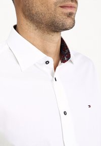 Uomo che indossa una camicia bianca con bottoni neri, un colletto interno a fantasia e un piccolo logo ricamato sul petto.