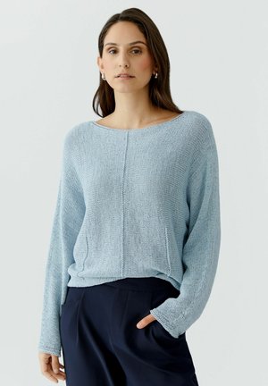 Femme aux longs cheveux noirs portant un pull en maille bleu clair et un pantalon foncé, debout devant un fond gris clair uni.