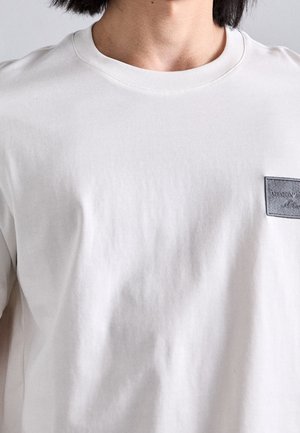 Persoon draagt een effen wit T-shirt met ronde hals en een klein rechthoekig zwart patch-logo op de linkerborst.