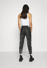 Jeans negros con una apariencia desgastada y texturizada, corte ajustado, costuras blancas y una etiqueta verde en la parte posterior. Se llevan con una camiseta sin mangas blanca y botas.