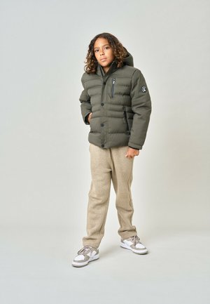 Enfant aux cheveux bouclés portant une doudoune verte, un pantalon en polaire beige et des baskets blanches, debout devant un fond uni clair.