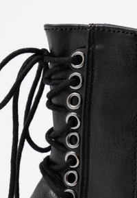 Stivaletto nero con lacci presenta una parte superiore in pelle sintetica testurizzata, occhielli argento e lacci neri, con un collo imbottito per il comfort.