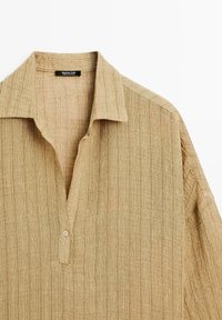 Camicia tan con texture, colletto, apertura con bottoni e maniche corte. Realizzata in tessuto leggero e intrecciato con righe verticali.