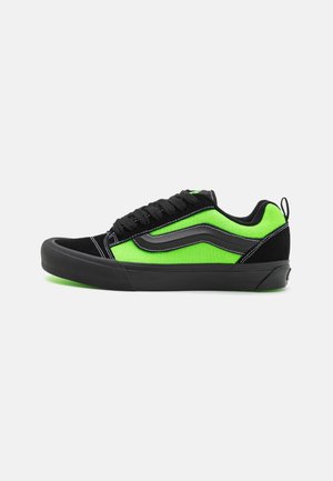 Lavtop sneaker med sort sål, sorte snørebånd og neongrønne sider med sort bølgedesign på hvid baggrund.