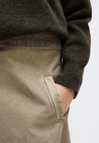 Χέρι σε τσέπη μπεζ παντελονιού, φοράει σκούρο πράσινο μαλακό πουλόβερ με μακριά μανίκια, εμφανιζόμενο από τη μέση έως το μέσο του μηρού, ουδέτερο φόντο.